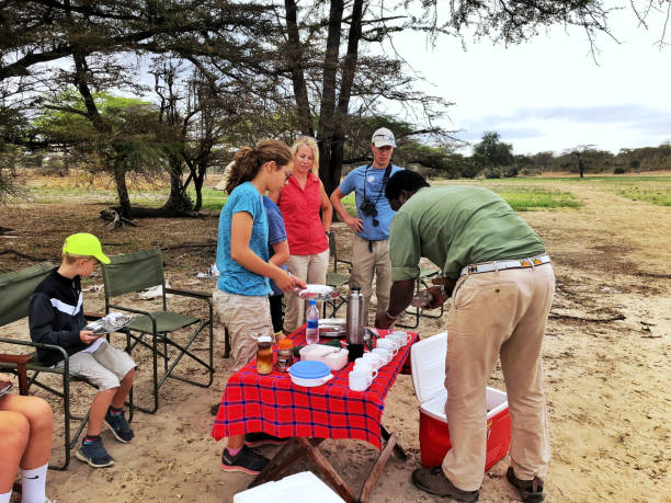 Family with children enjoying breakfast in front of a safari Land Rover near a lake during a morning safari in Selous Game Reserve, Tanzania